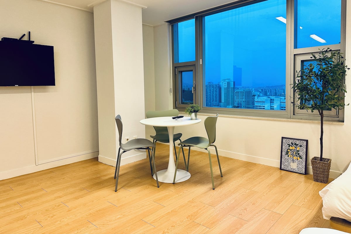 Une pièce moderne dotée d’une grande fenêtre, d’une table ronde avec des chaises, d’une plante en pot et d’un téléviseur à écran plat fixé au mur.