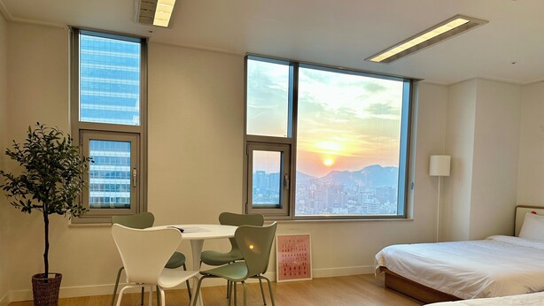 Habitación de hotel con cama, una mesa de comedor con sillas, una planta en maceta y ventanas amplias con vistas a la ciudad.