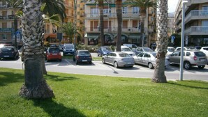A street with parked cars and palm trees in the foreground. In the background, there are multi-story buildings with balconies and a sign that reads "Pizzeria." - Hotel Giordana (Loano)