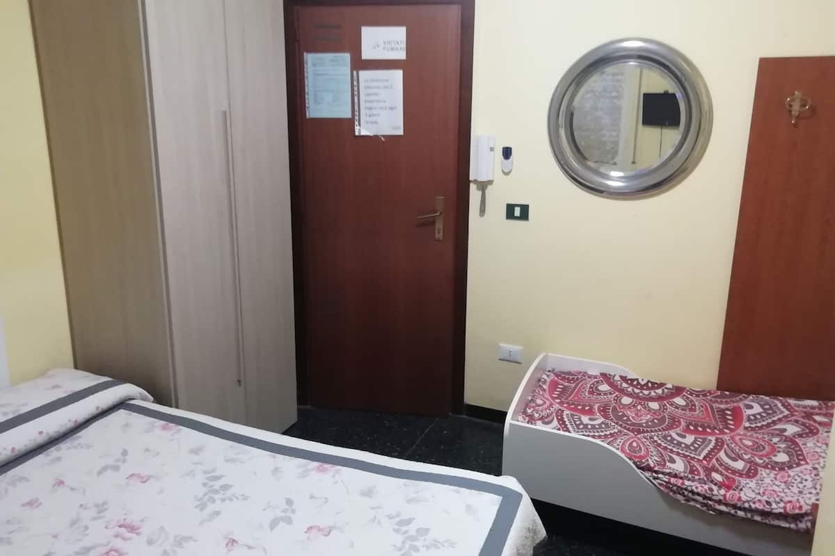 Une chambre comprenant un lit, une armoire, une porte où sont affichées des avis, un miroir fixé au mur et une couvre-lit à motifs.