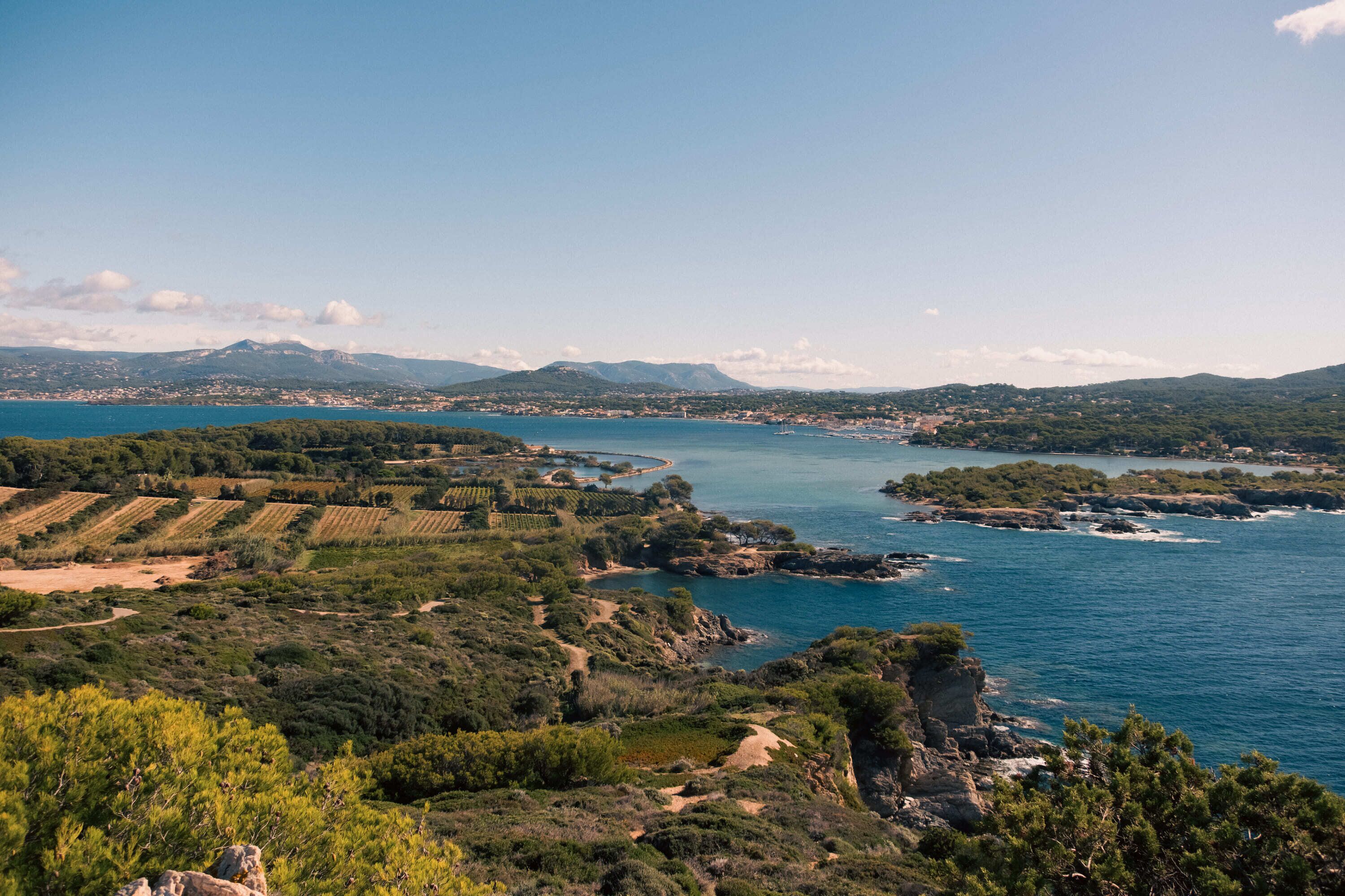 A coastal landscape with vineyards, rocky terrain, and a view of the sea and distant mountains.