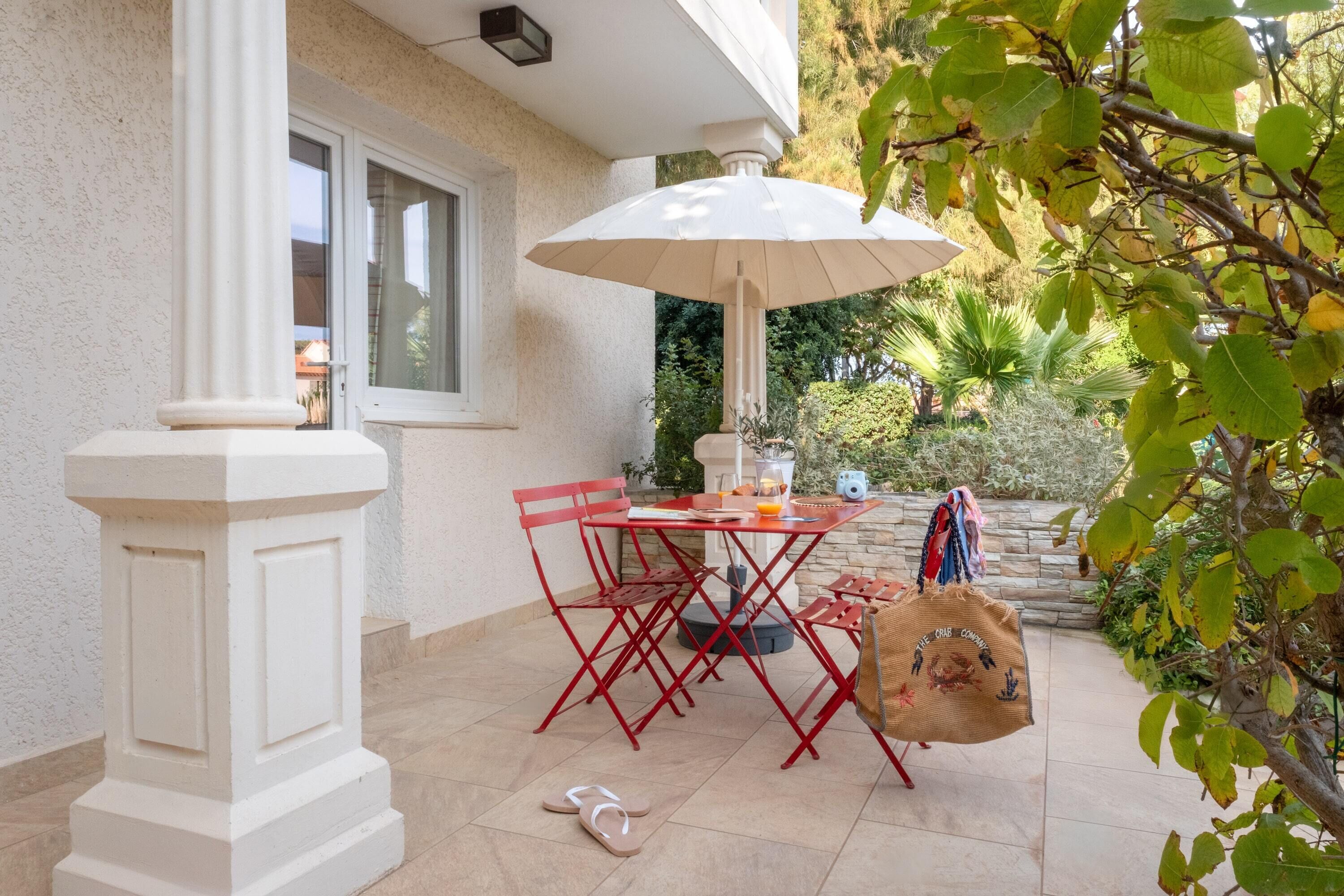 An outdoor patio with a white column, red metal chairs, a table with a white umbrella, and a stone wall in the background.