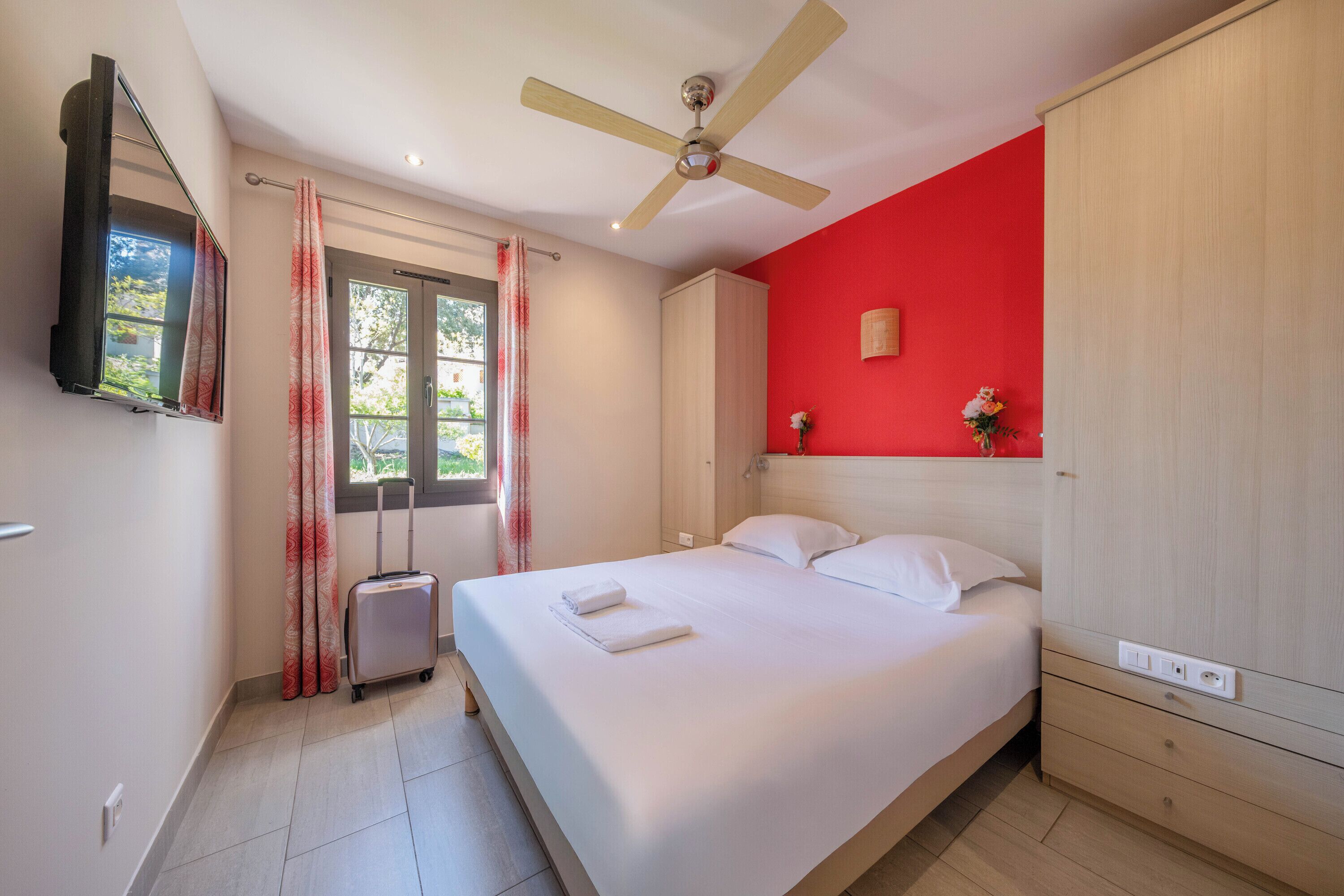 Une chambre à coucher avec un lit, un ventilateur de plafond, une fenêtre avec des rideaux et un mur rouge.