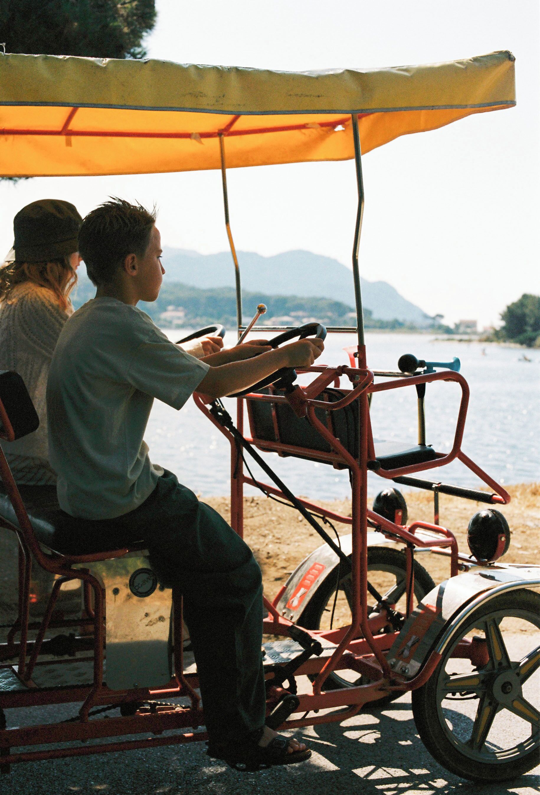 A man driving a small, open vehicle with a canopy, parked near a body of water with mountains in the background.