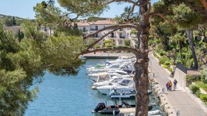 A marina with boats docked along a waterfront, a pedestrian path beside the water, and buildings in the background. - Appartements de l'Hélios - Îles des Embiez (Six-Fours-les-Plages)