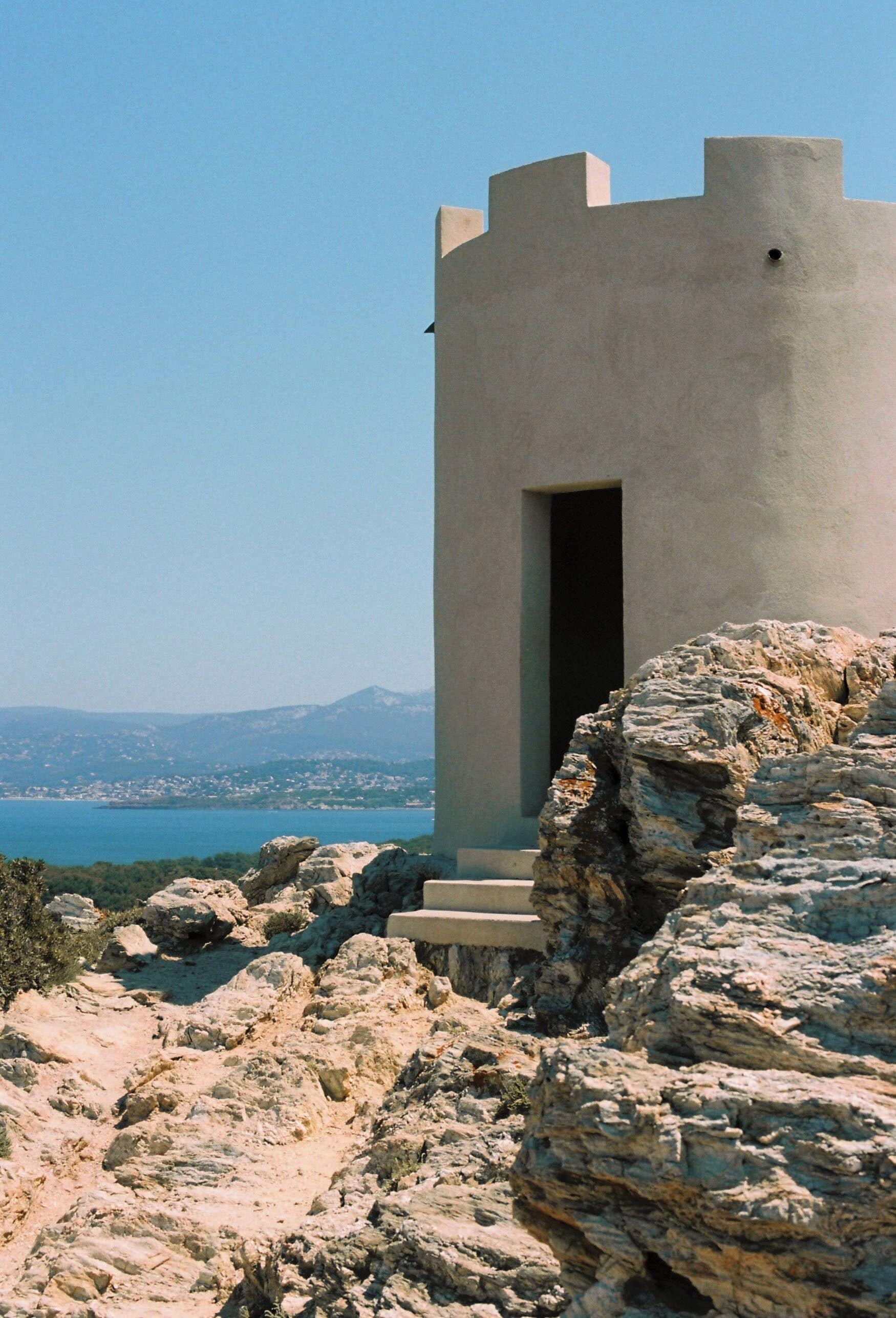 A modern building with a tower-like structure, a doorway, and a rocky landscape with a distant view of a body of water and mountains.