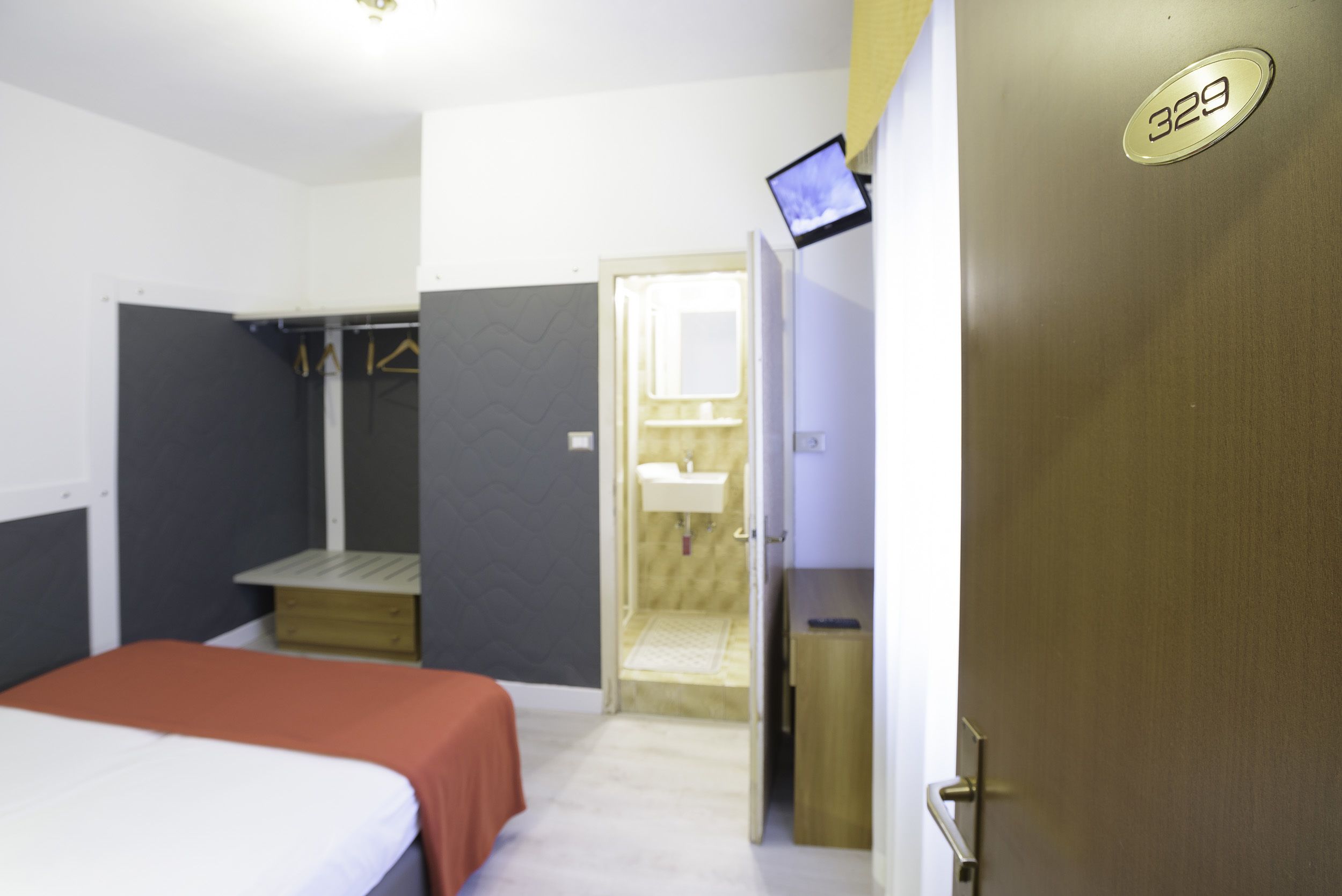 A bedroom with a bed, a desk, a wardrobe, a bathroom with a sink, and a door with a number plate.