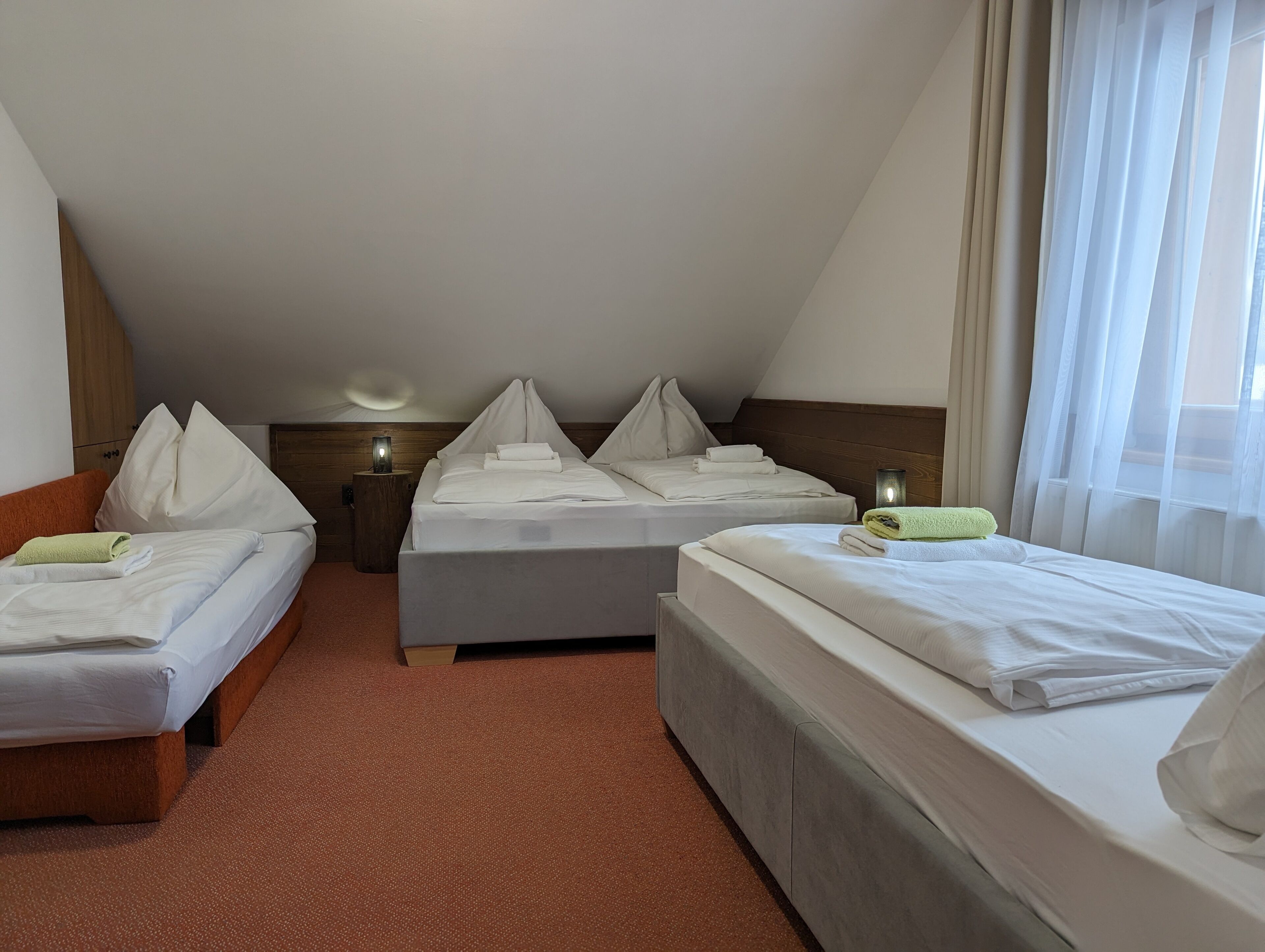 Habitación de hotel con dos camas individuales, cabecero de madera, una mesita con lámpara y una ventana con cortinas de gasa.