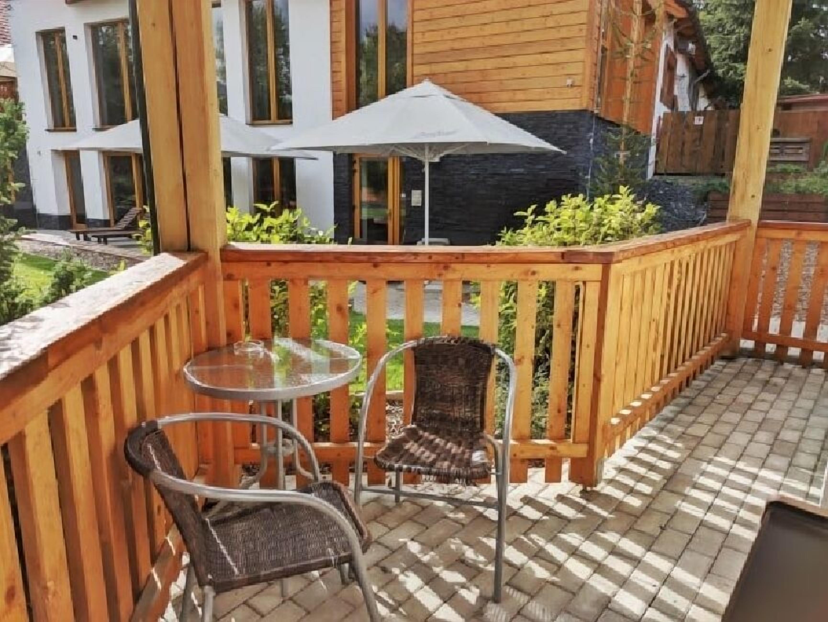 A wooden deck with a glass table and two wicker chairs, a wooden fence, and a building in the background.