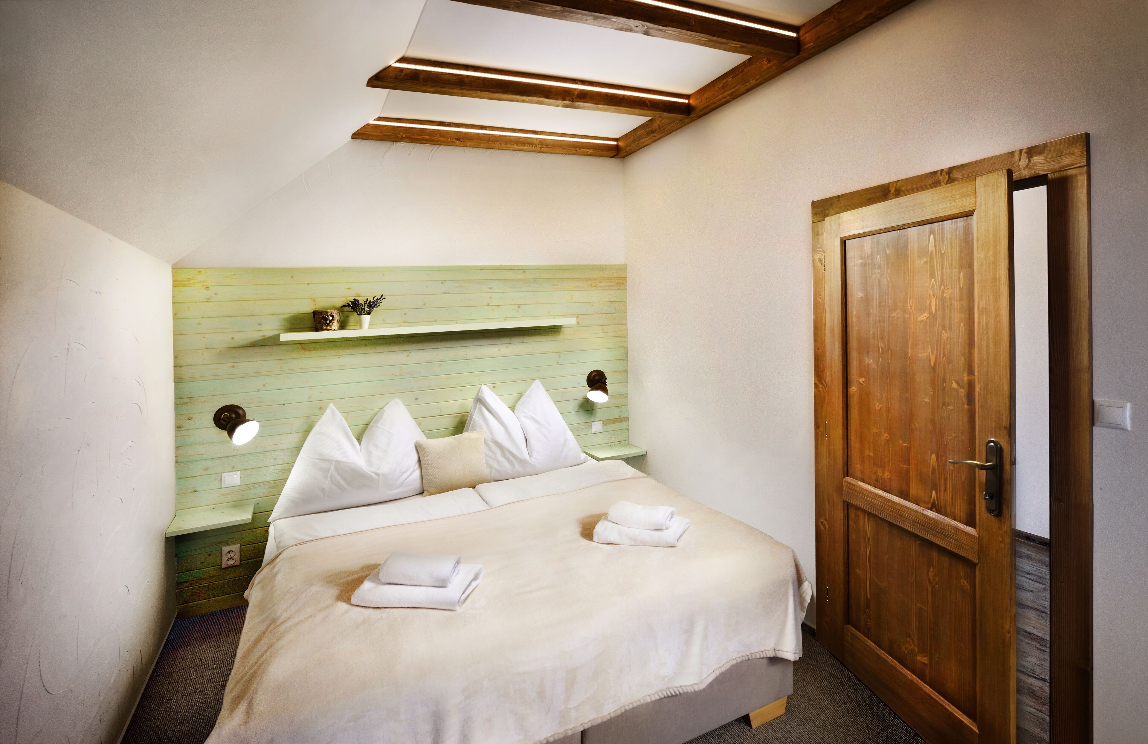 A bedroom with a bed, pillows, a nightstand, a shelf with decorative items, and a wooden door.