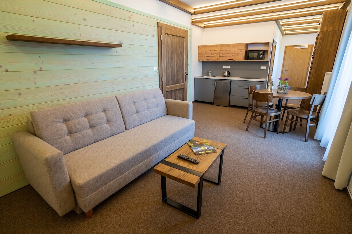 A compact living space with a sofa, a small coffee table, a dining area with chairs, a kitchen with cabinets and appliances, and a wooden door.