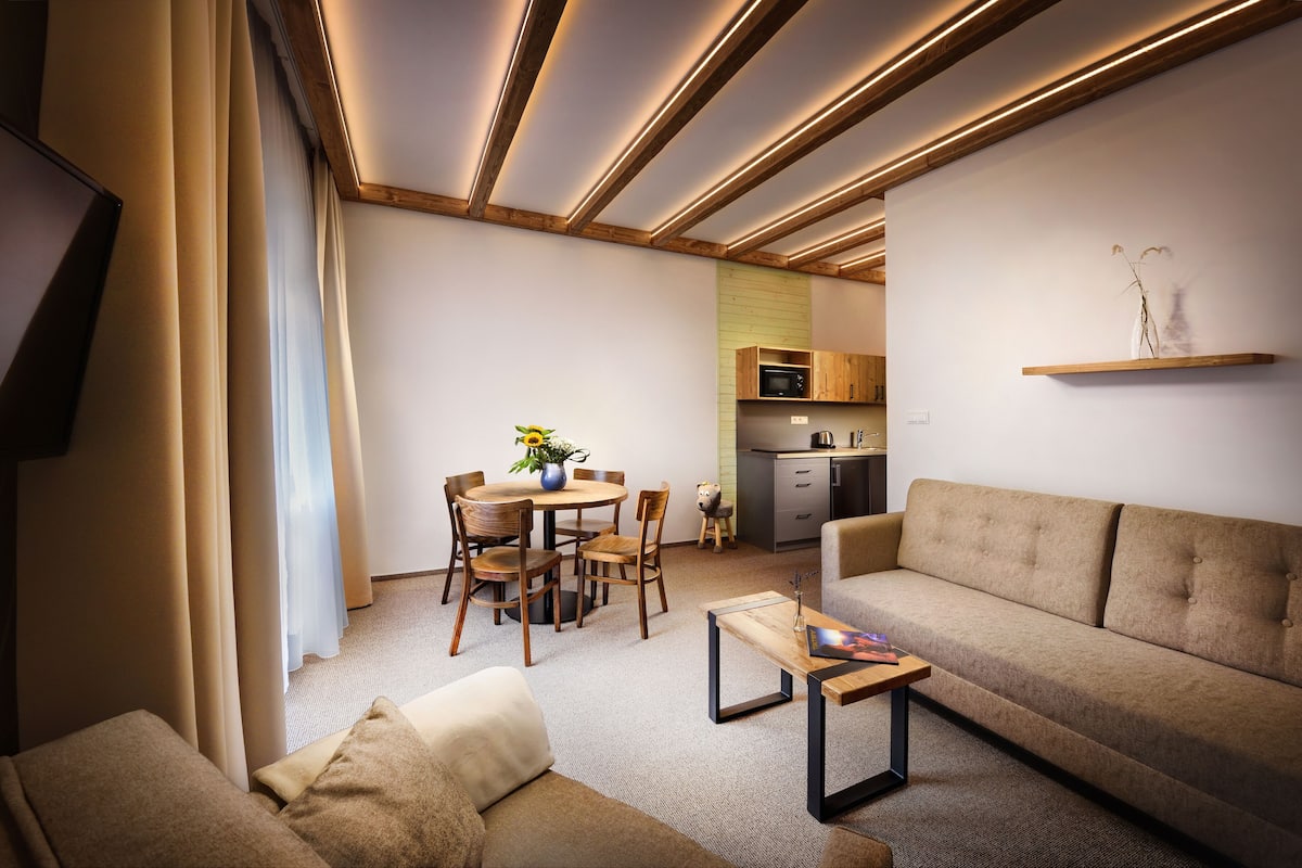 A modern living room with a sofa, a coffee table, and a dining area with a table and chairs. There is a kitchen area with a microwave and cabinets.