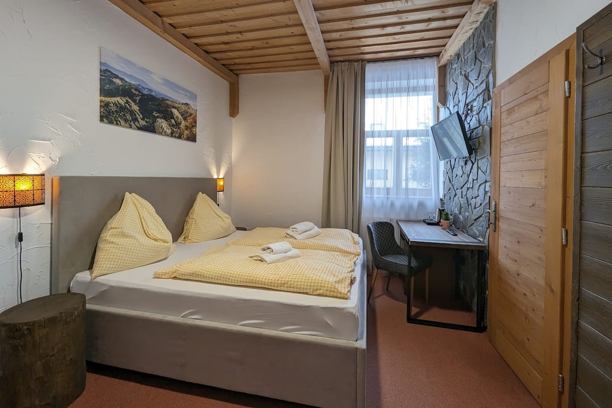 A bedroom with a bed, a desk, a chair, a lamp, a painting, and a stone wall.
