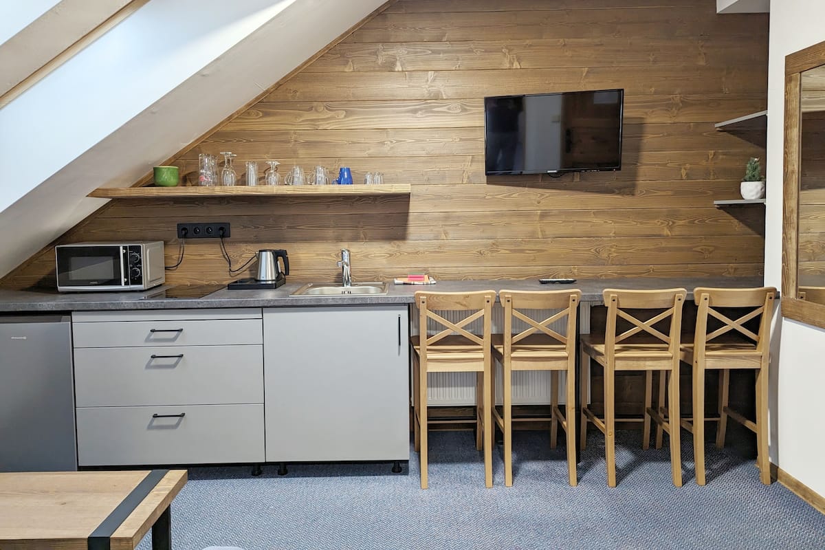 A compact kitchen with a wooden wall, a flat-screen TV, a microwave, and a sink. There are four wooden chairs and a small table.