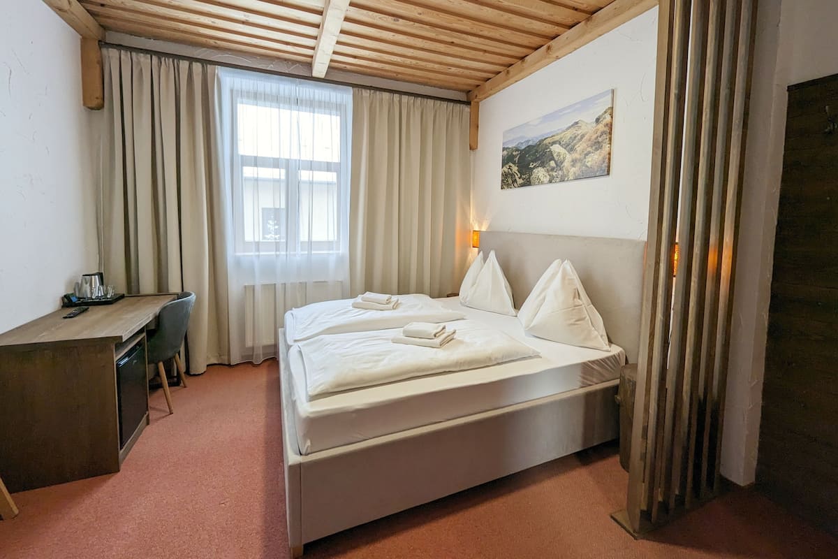 A hotel room with a wooden ceiling, a bed with white linens, a desk with a chair, a window with curtains, and a painting on the wall.