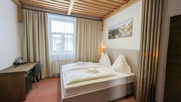 Habitación de hotel con techo de madera, cama con ropa blanca, escritorio con silla, ventana con cortinas y un cuadro en la pared.