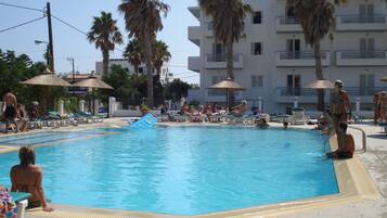 Una piscina con sillones de descanso y sombrillas, rodeada de palmeras y un edificio de varios pisos.