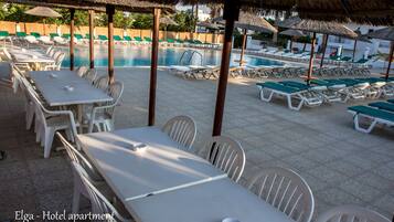Zona de piscina de hotel con sillones y mesas bajo sombrillas de paja.