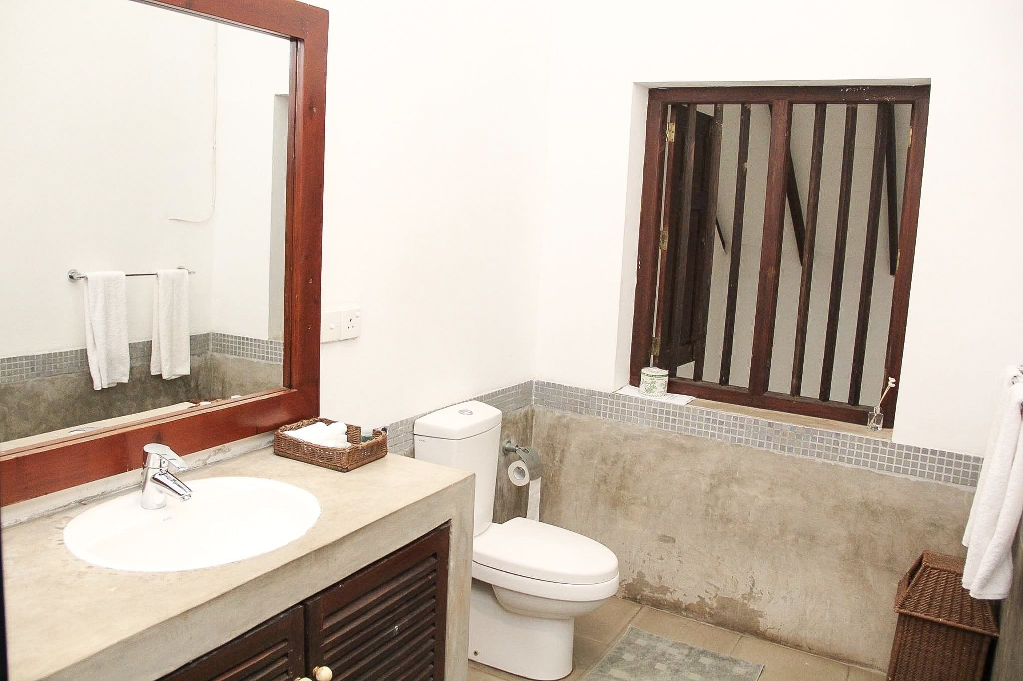 Un baño con un lavamanos blanco, un espejo, un inodoro y una ventana con marco de madera.