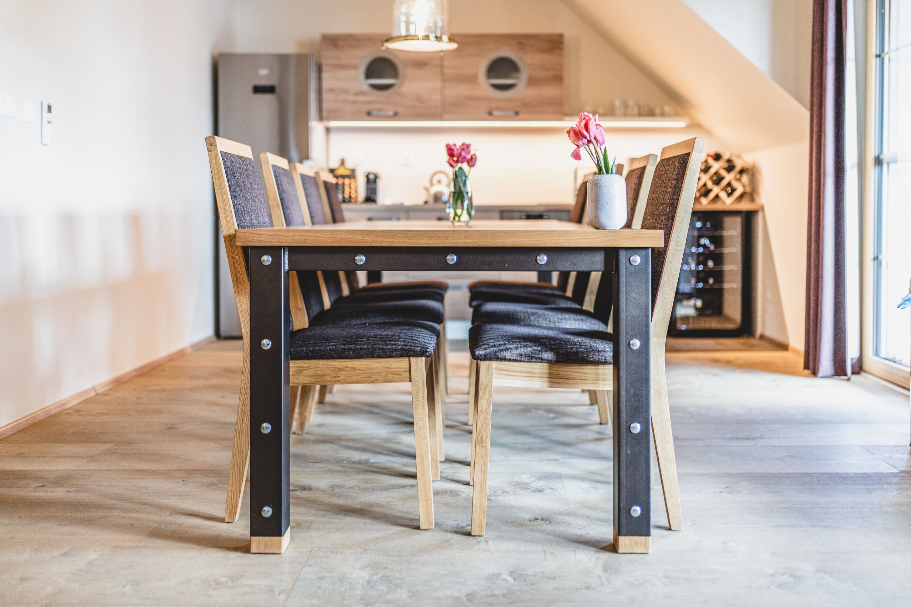 A dining area with a wooden table and chairs, a vase with flowers, and a kitchen in the background.