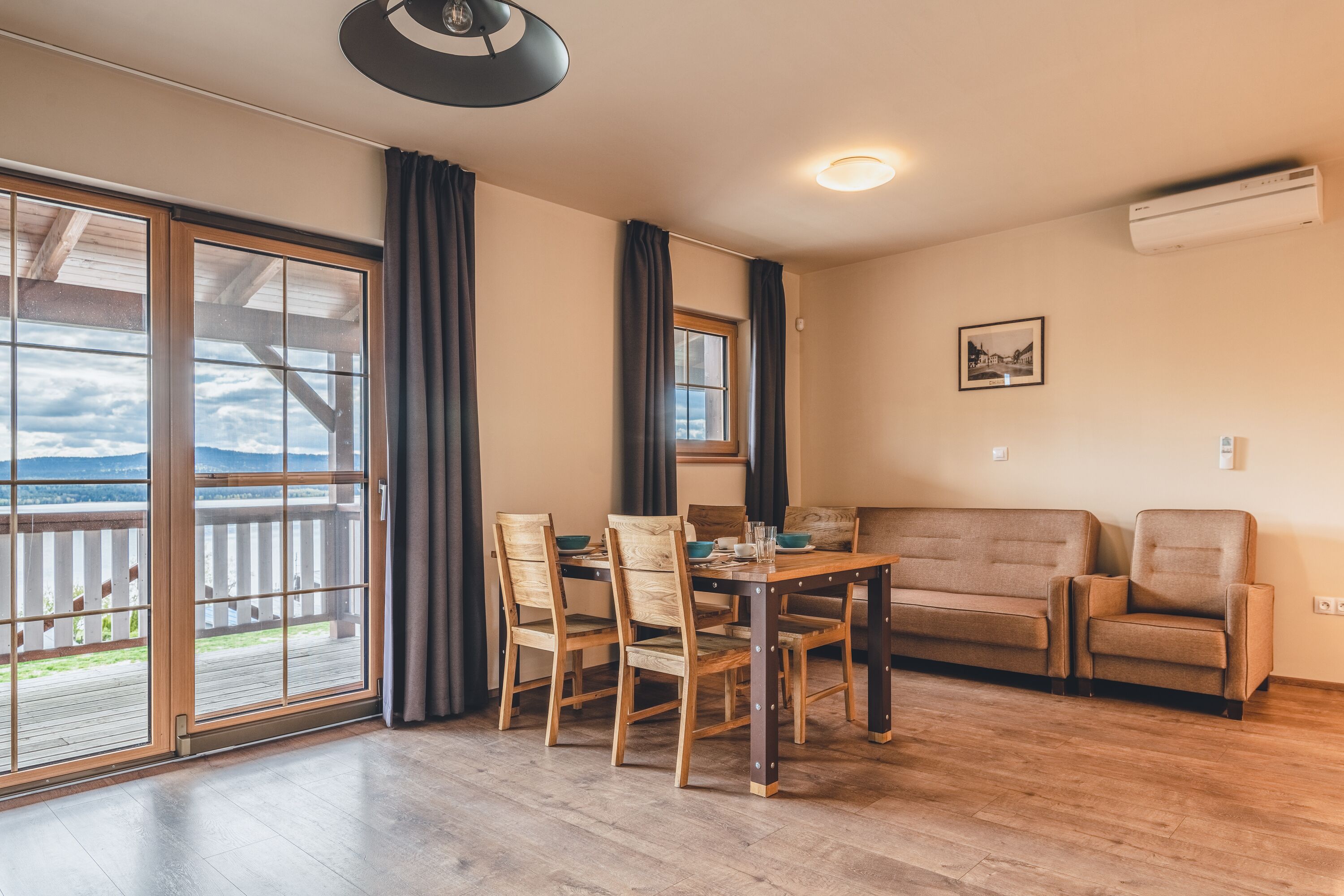 A room with a wooden dining table, four chairs, a sofa, and a large sliding glass door leading to a balcony.