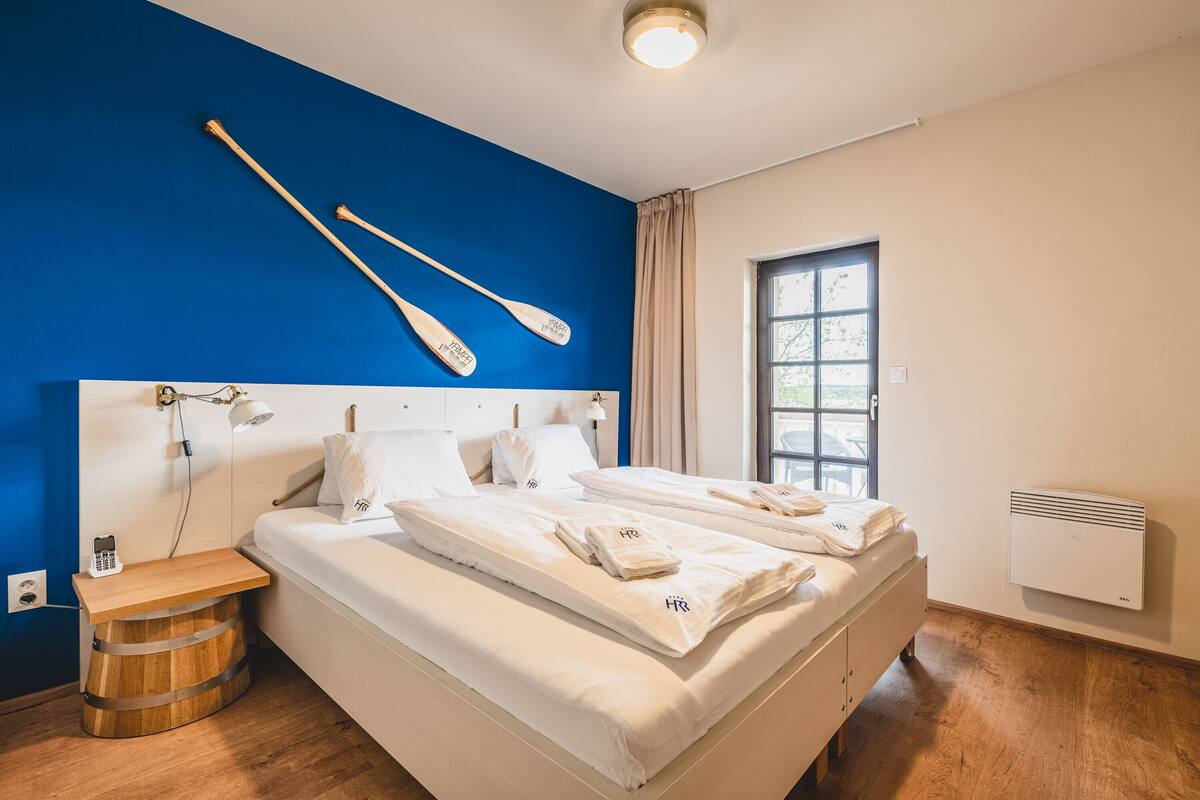 Ein Doppelbett mit weißer Bettwäsche, ein Nachttisch aus Holz mit einem Telefon und ein kleiner Holzbärgel.
