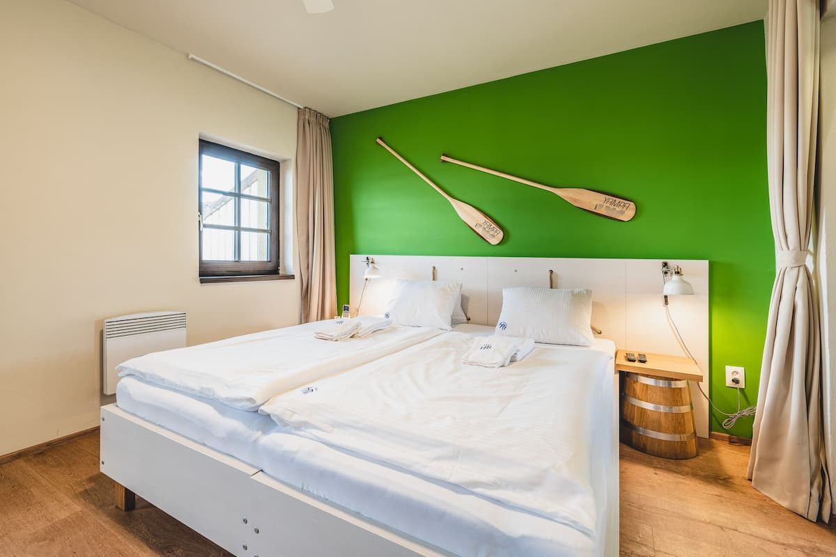 Ein Doppelbett mit weißen Leinen, ein Holz-Kopfteil, ein Fenster mit Vorhängen, ein an der Wand befestigtes Holzbrett und ein Nachttisch mit einer Lampe.