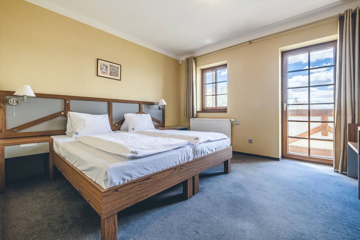 Ein Hotelzimmer mit einem hölzernen Bett, zwei Nachttischen mit Lampen, einem Fenster mit Vorhängen und einer Heizung.