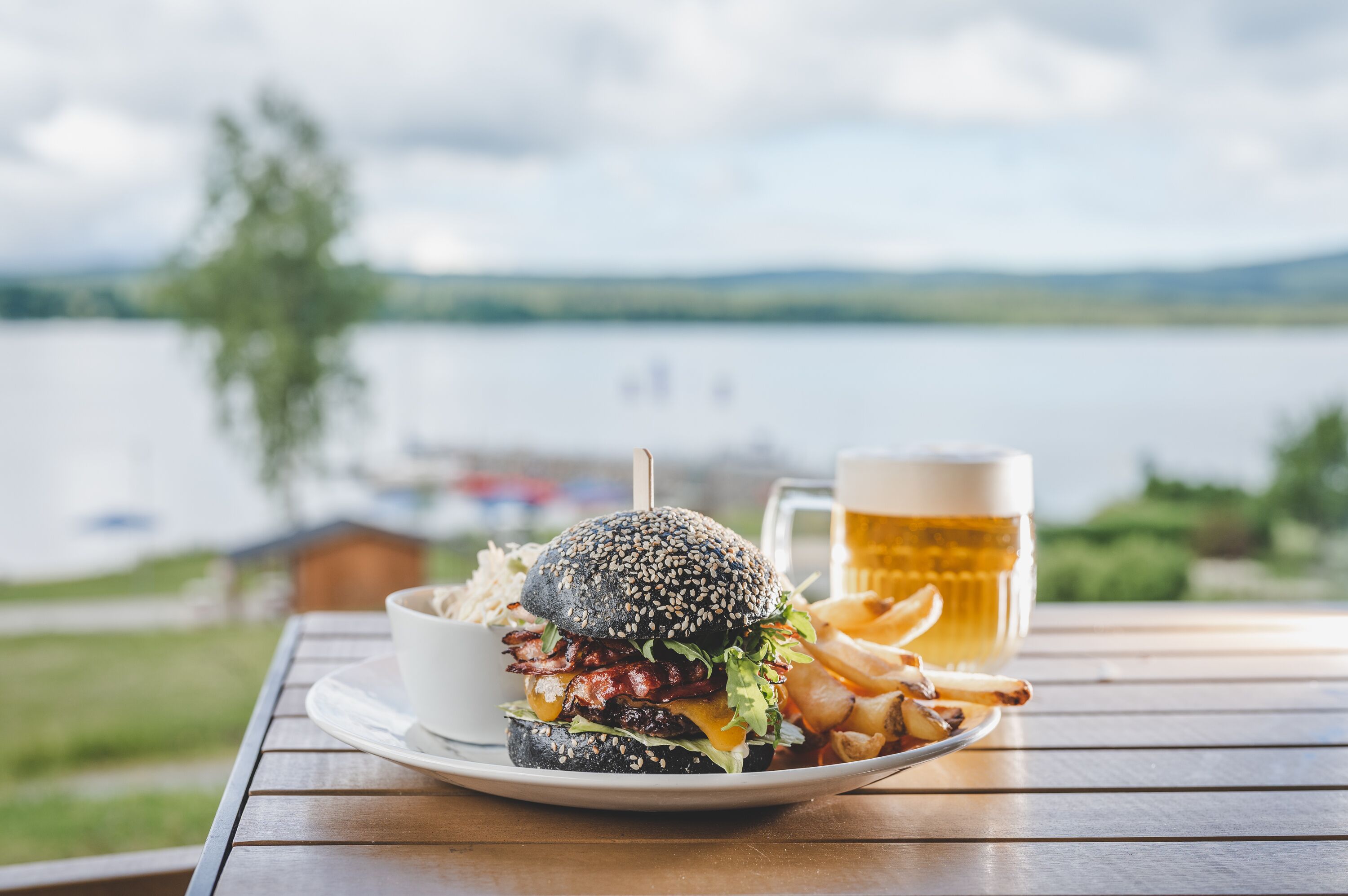 A meal of a burger, fries, and a beer on a wooden table with a view of a lake and distant buildings.