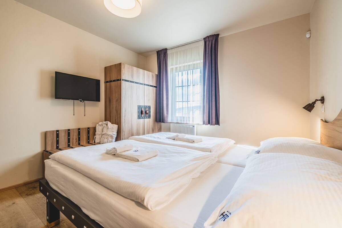 Ein Hotelzimmer mit zwei Betten, einem an der Wand montierten Fernseher, einem Holzschrank und einem Fenster mit Vorhängen.