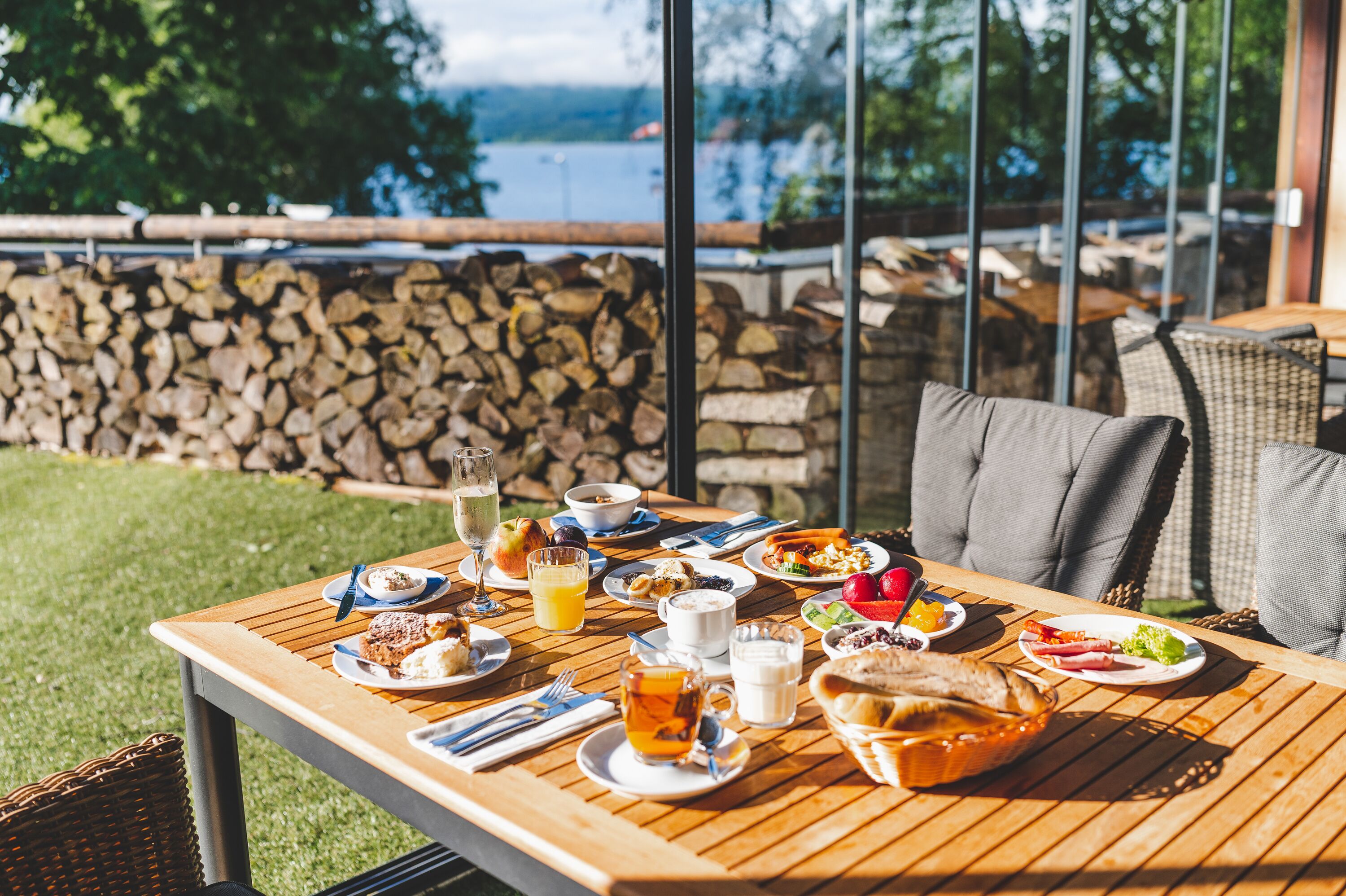 Una mesa de comedor al aire libre con diferentes alimentos para el desayuno, una cesta de mimbre y vistas a un lago y árboles.