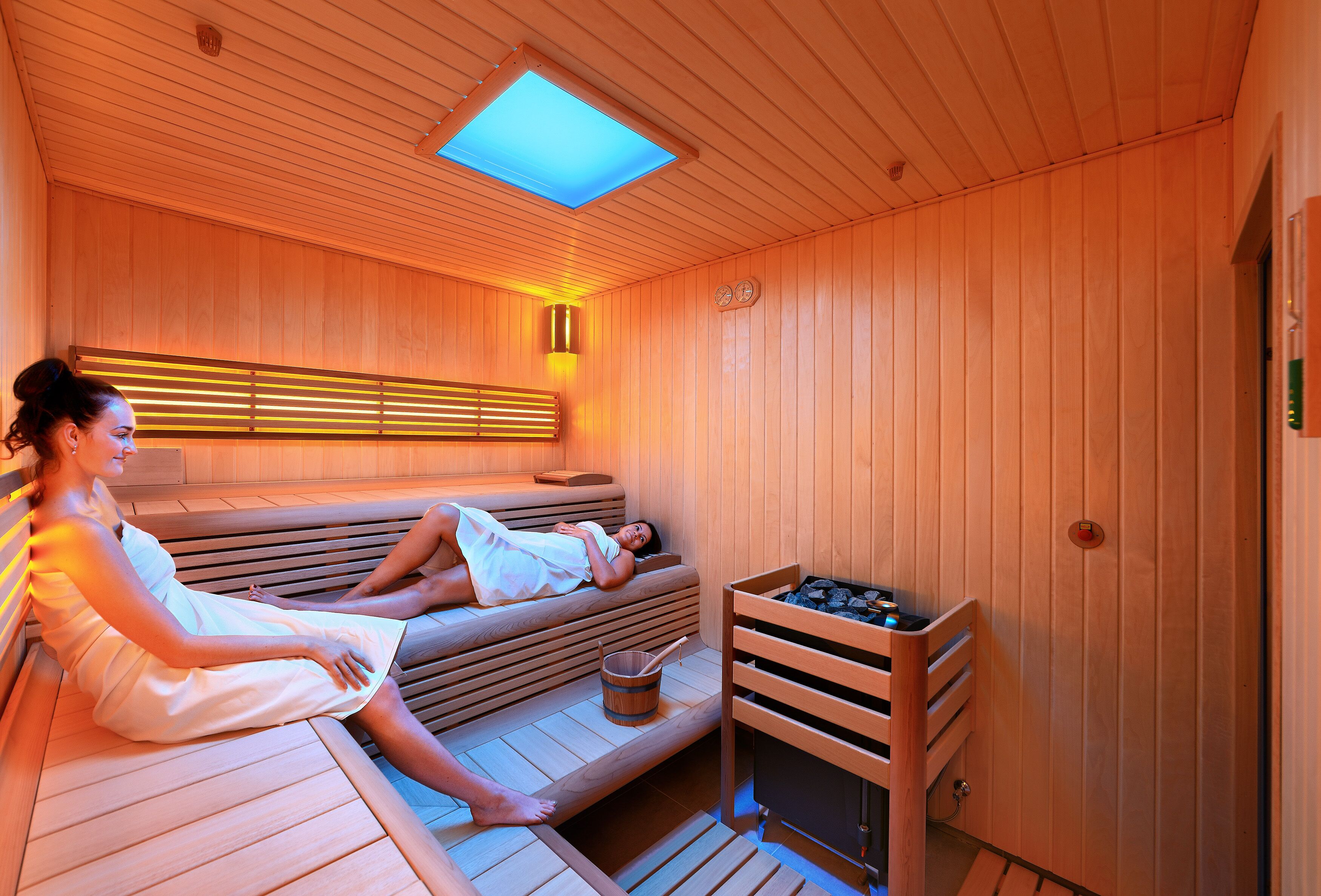 Un sauna con bancos de madera y un asiento. Hay un balde con una cuchara y un calentador en el banco.