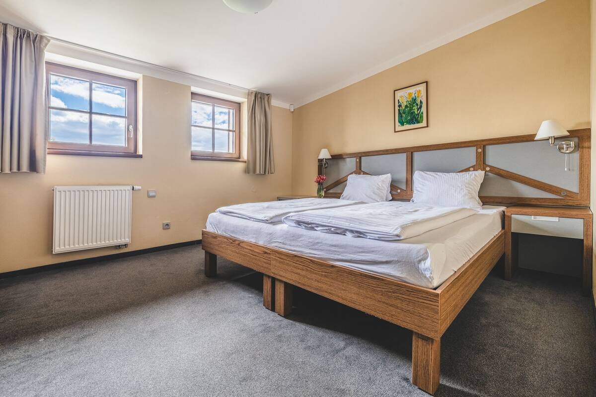 Ein Schlafzimmer mit einem Holzbett, zwei Nachttischen, einem Heizkörper und zwei mit Vorhängen versehenen Fenstern.