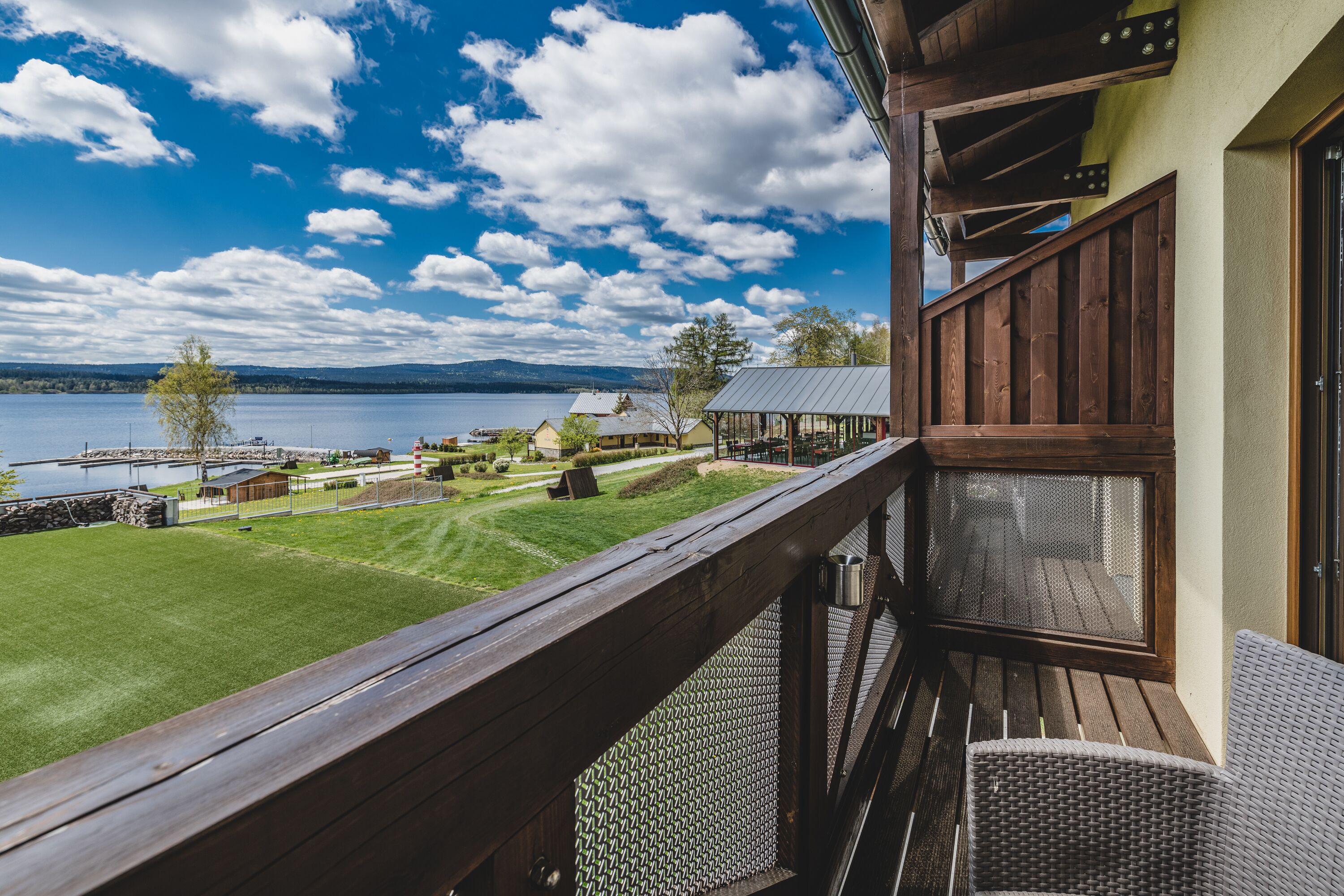 A balcony with a view of a lake, a grassy area, and a building in the background.