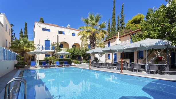 Una piscina con sillones de descanso y sombrillas, rodeada de edificios con puertas y ventanas azules.