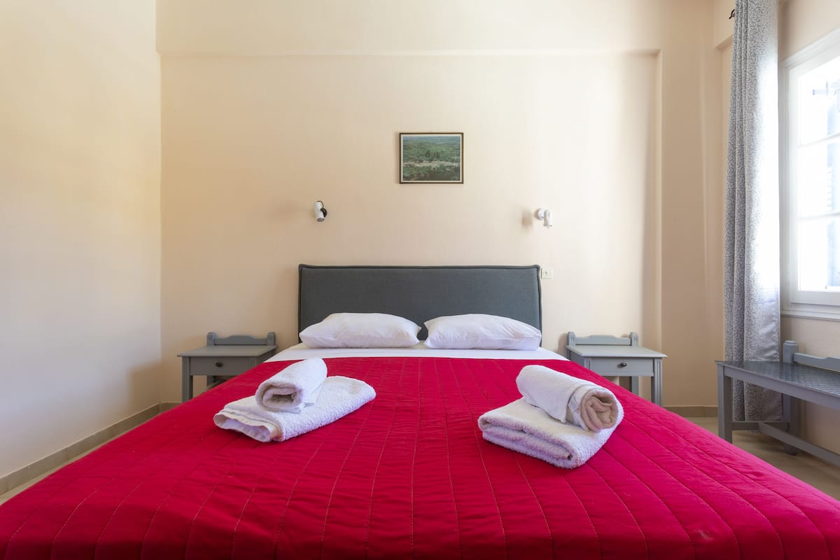 A hotel room with a large bed, two bedside tables, a nightstand, a window with curtains, and a painting on the wall.