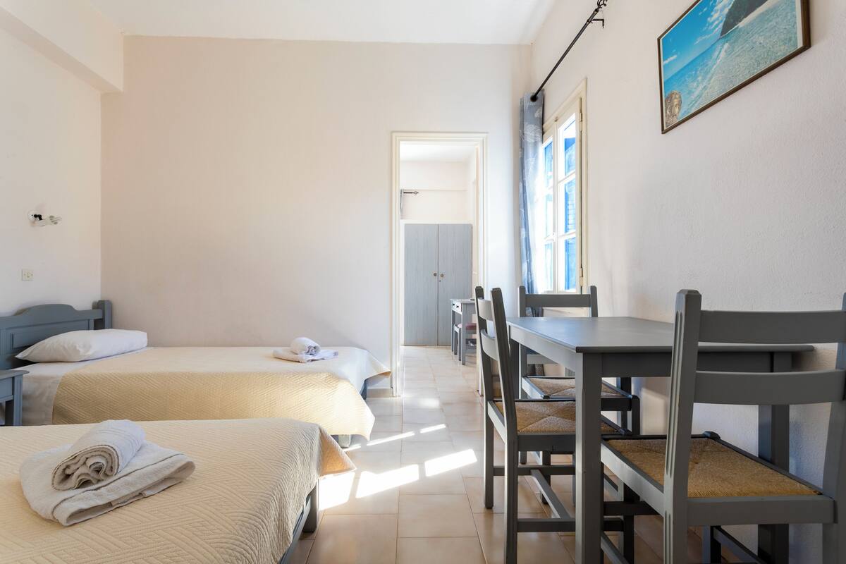 A hotel room with two beds, a dining table with chairs, and a view of the sea through the window.