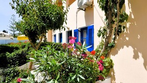 Un edificio con contraventanas azules y pared blanca, rodeado de abundante follaje y plantas en flor.