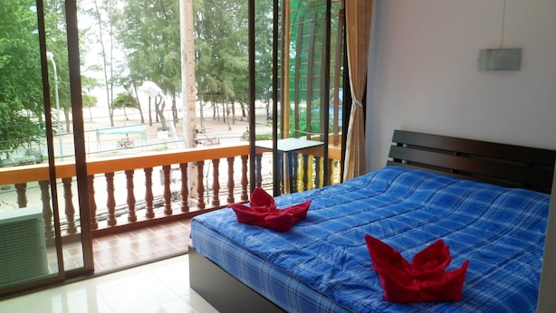 A hotel room with a large window, a bed with blue bedding, and a balcony with a view of the street and trees. - Onn Inn at Cha-am (Cha-am)