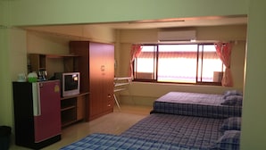 A compact room with a kitchenette, a bed with a checkered duvet, a window with curtains, and a built-in air conditioning unit. - Onn Inn at Cha-am (Cha-am)