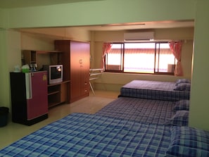 A compact room with a kitchenette, a bed with a checkered duvet, a window with curtains, and a built-in air conditioning unit.