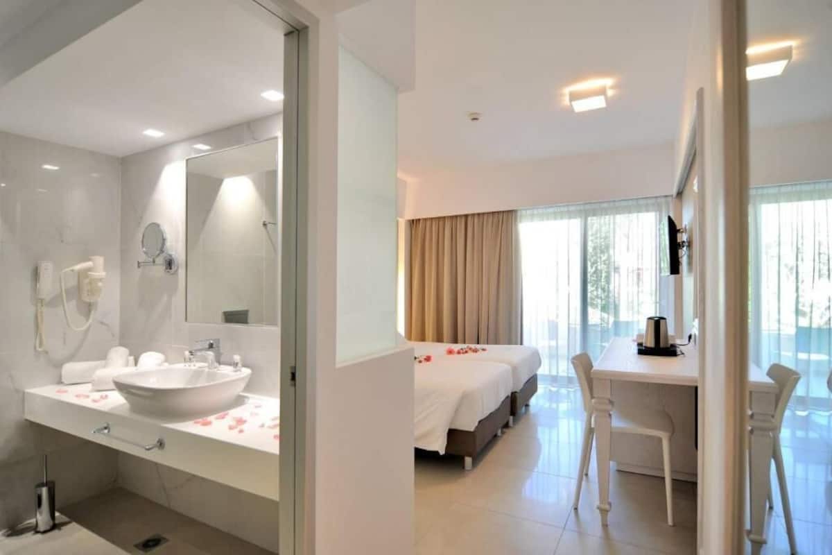 Una habitación de hotel moderna con un baño amplio, una cama con pétalos de rosa, un escritorio y un ventanal grande.