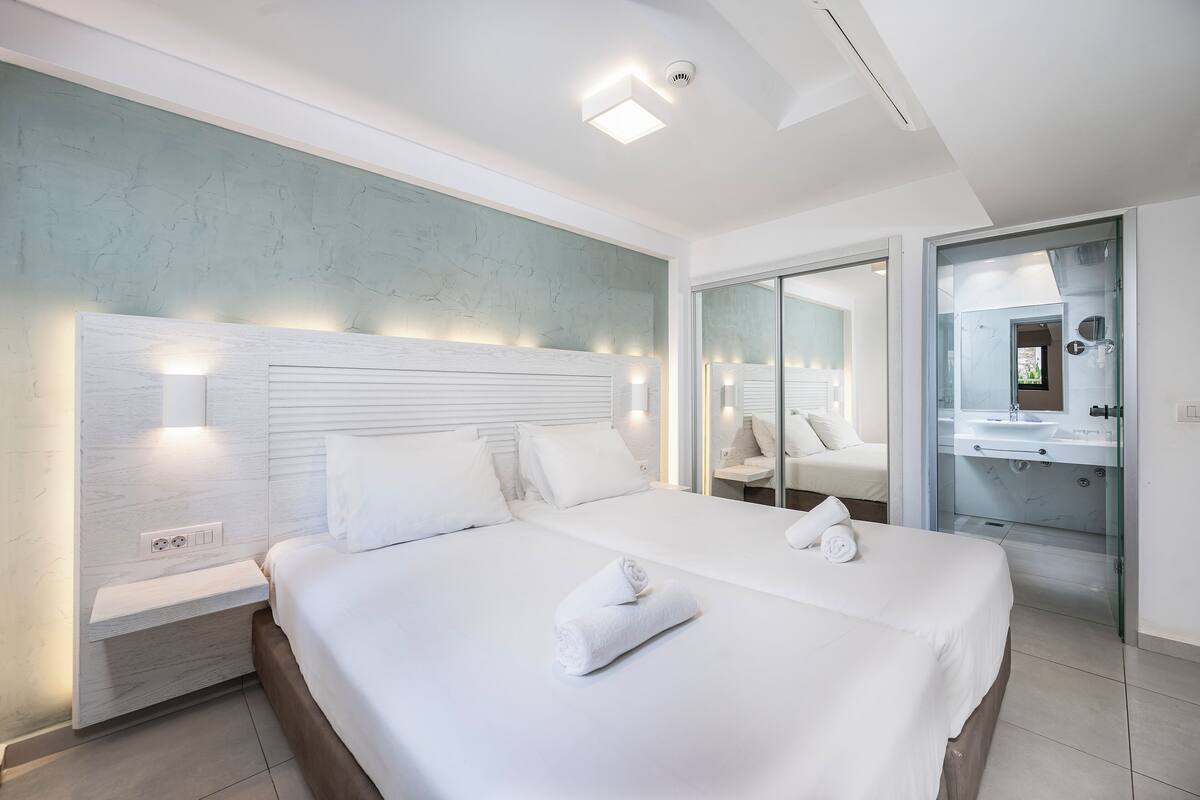 Una habitación de hotel moderna con una cama grande, mesitas de noche, un baño con paredes de vidrio y vistas a la ciudad.