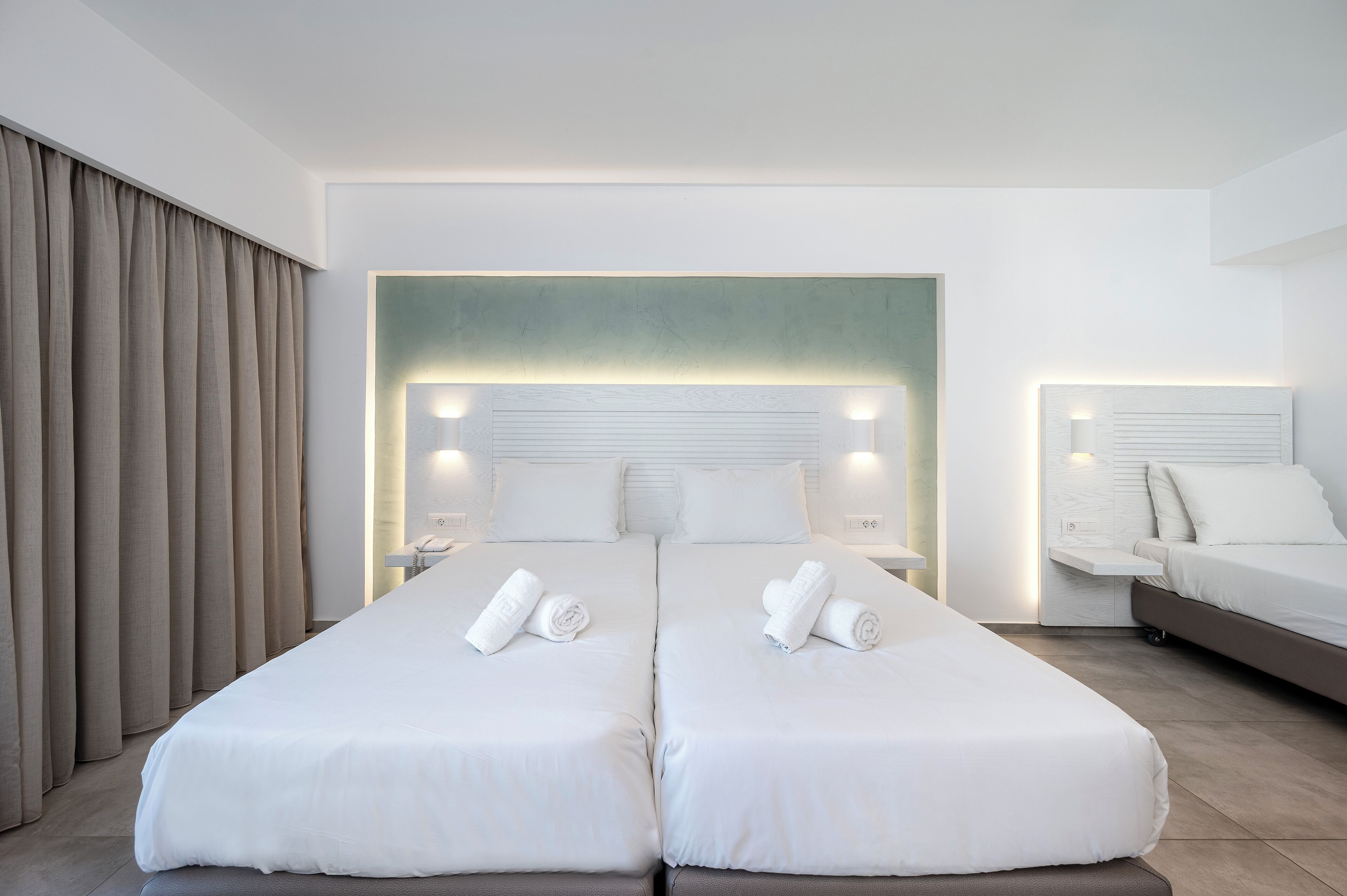 A modern hotel room with two single beds, a sofa, a large window with curtains, and a wall-mounted light fixture.
