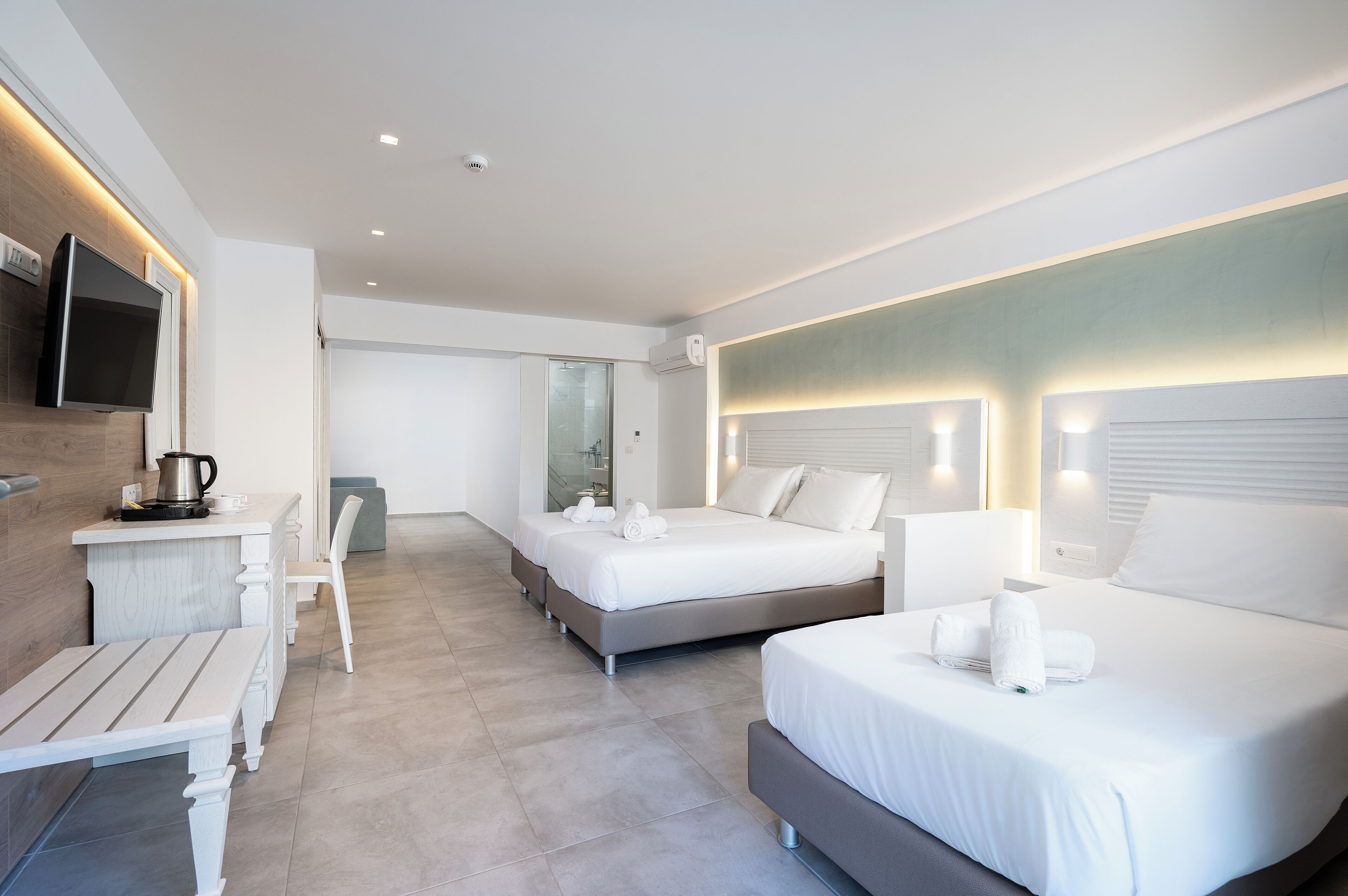 A modern hotel room with a large bed, a smaller bed, a desk with a kettle, and a TV mounted on the wall.