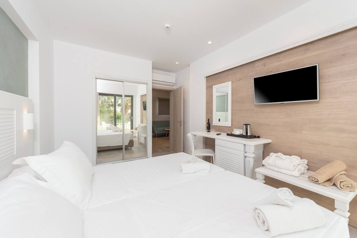 Una habitación de hotel moderna con una cama grande, un televisor de pantalla plana montado en la pared, un tocador con espejo y una pequeña zona de estar con una mesa y sillas.