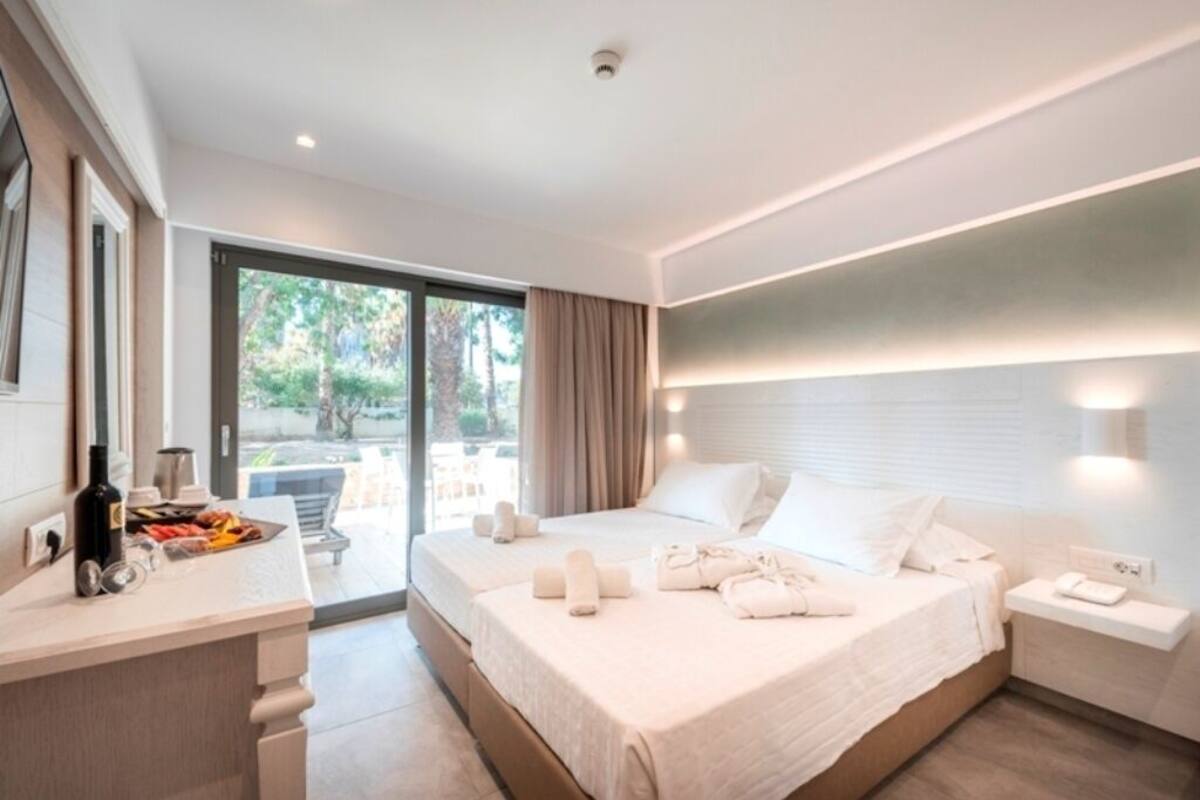 Una habitación de hotel moderna con una cama grande, una mesita de noche, un escritorio con una botella y vasos, y vistas al exterior.