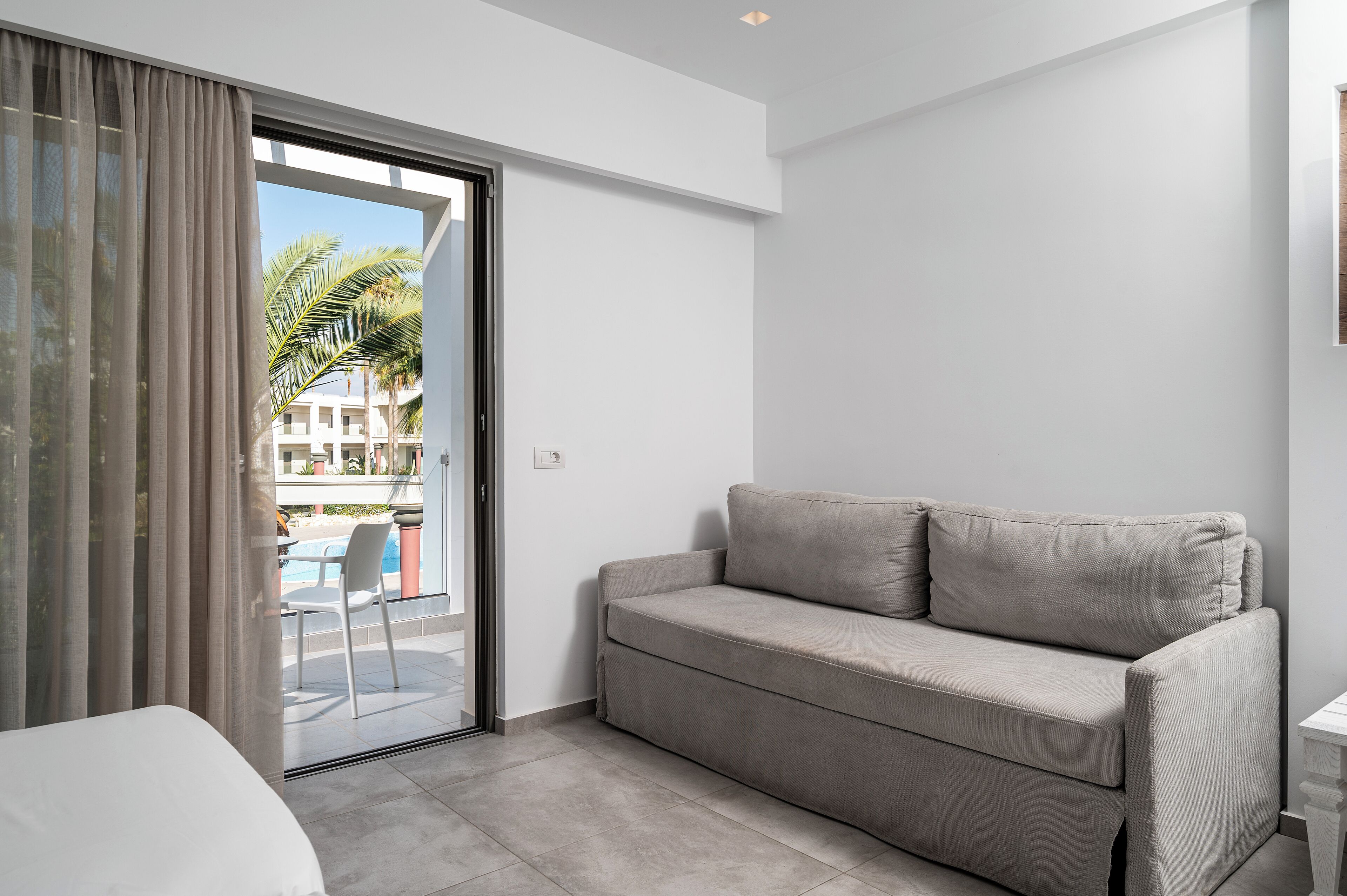 Een moderne kamer met een grijze bank, een glazen deur naar een balkon en een witte muur.