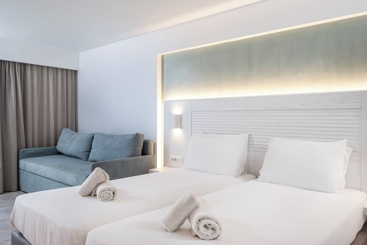Habitación de hotel con dos camas, un sofá y un amplio ventanal con cortinas.