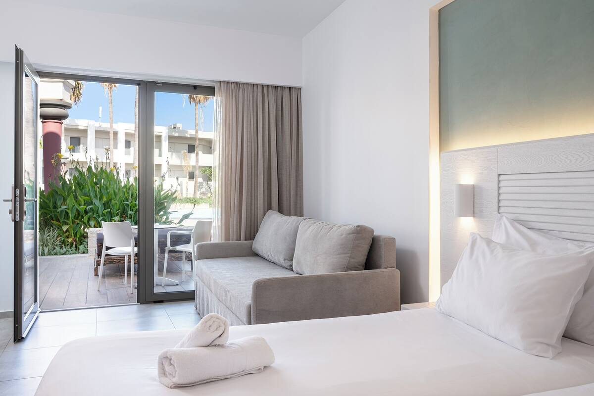 Habitación de hotel con cama, sofá y puerta corrediza de vidrio que da a un área exterior con patio y plantas.